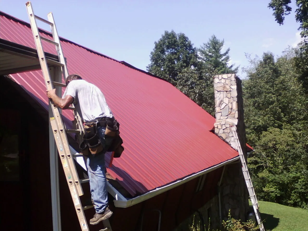 Roofer on a ladder beside a red metal roof and stone chimney — Soffit & Fascia Repair by Roofing Brothers in Point Of Rocks